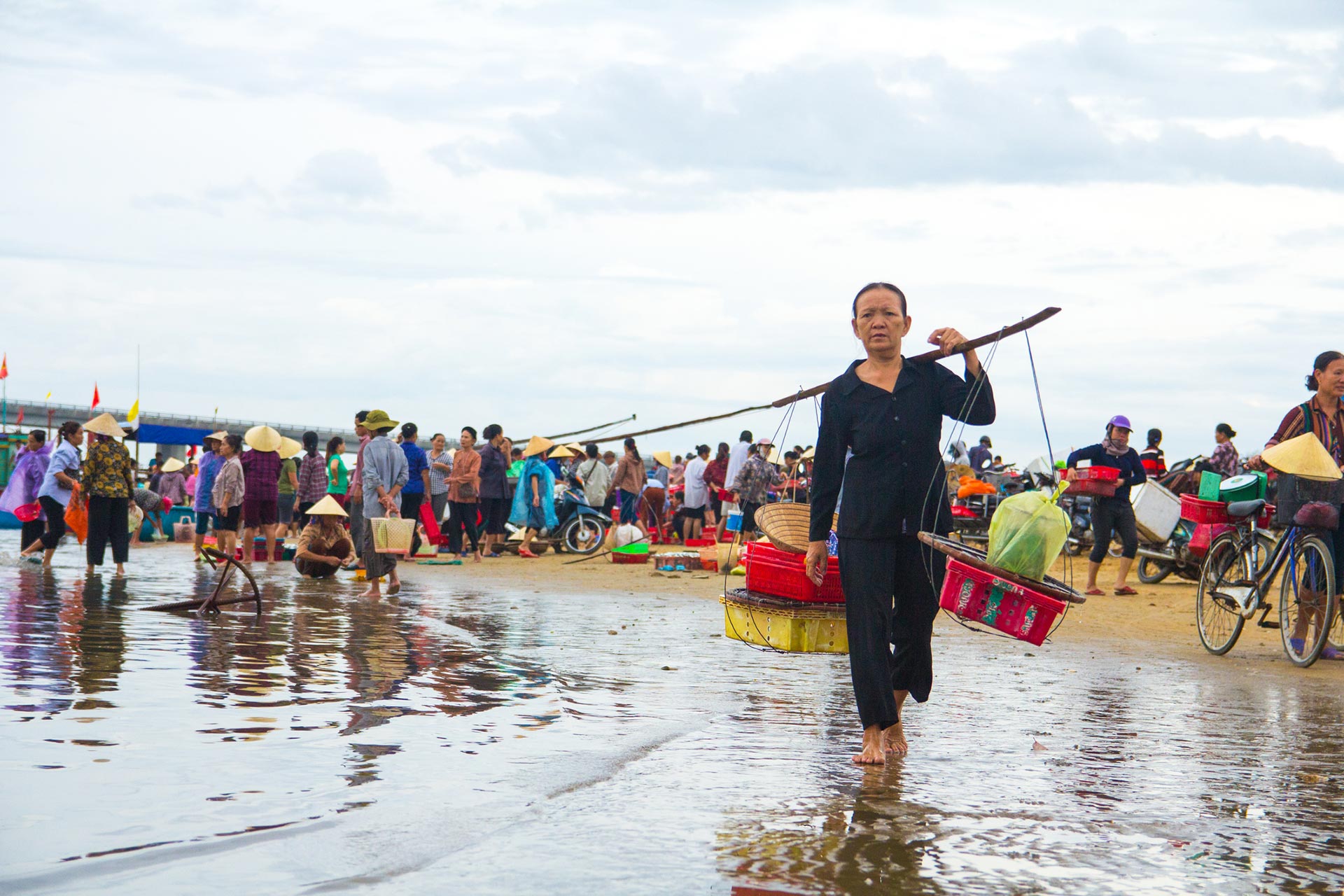 Fra stranden i Hà Tĩnh. En eldre vietnamesisk kvinne bærer plastkurver i forgrunnen. Bak henne er det en folkemengde på et fiskemarked. 