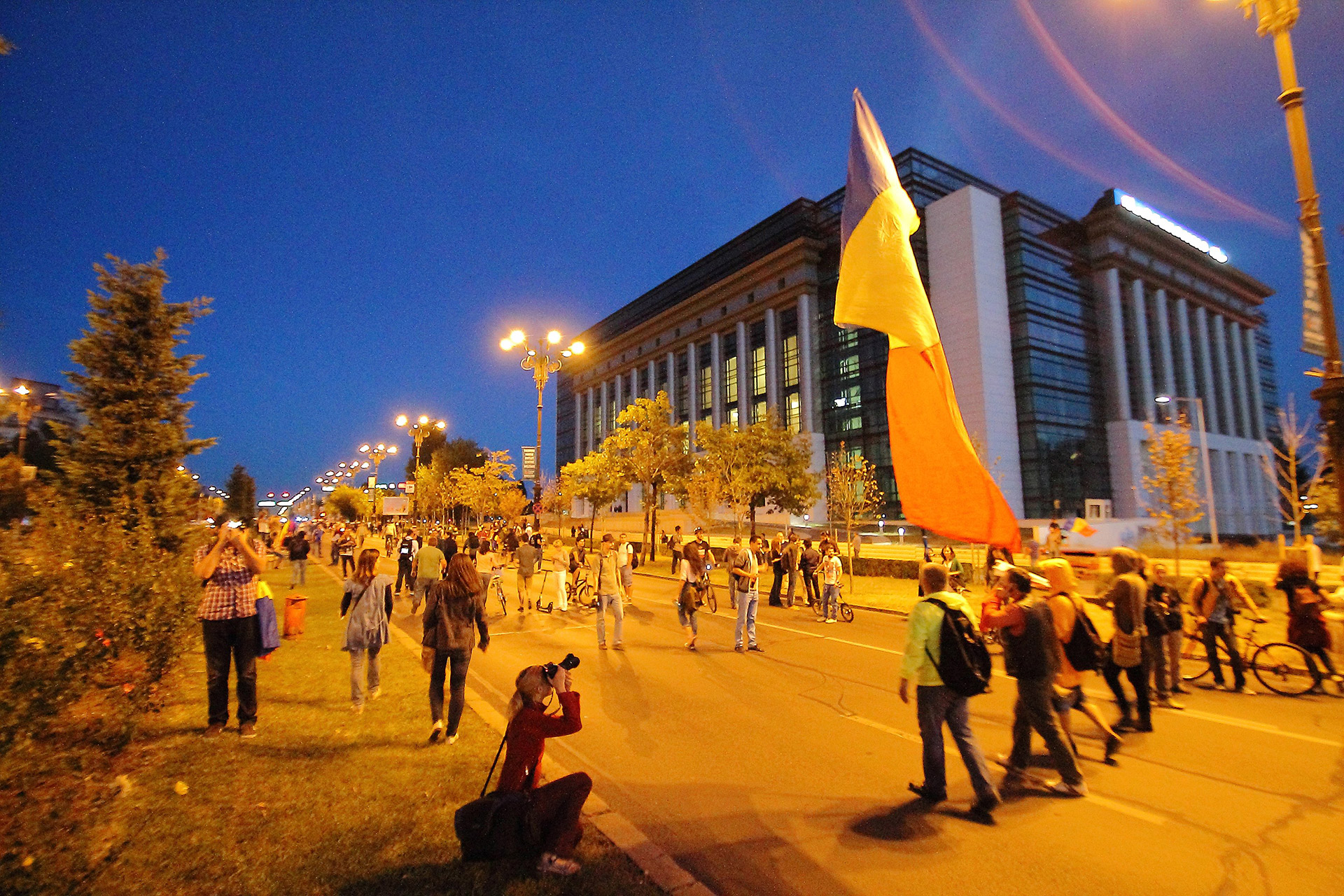 emonstranter går på veien etter solnedgang, 15. september 2013. En har et stort rumensk flagg, flere tar bilder. I bakgrunnen ser man det rumenske nasjonalbiblioteket.