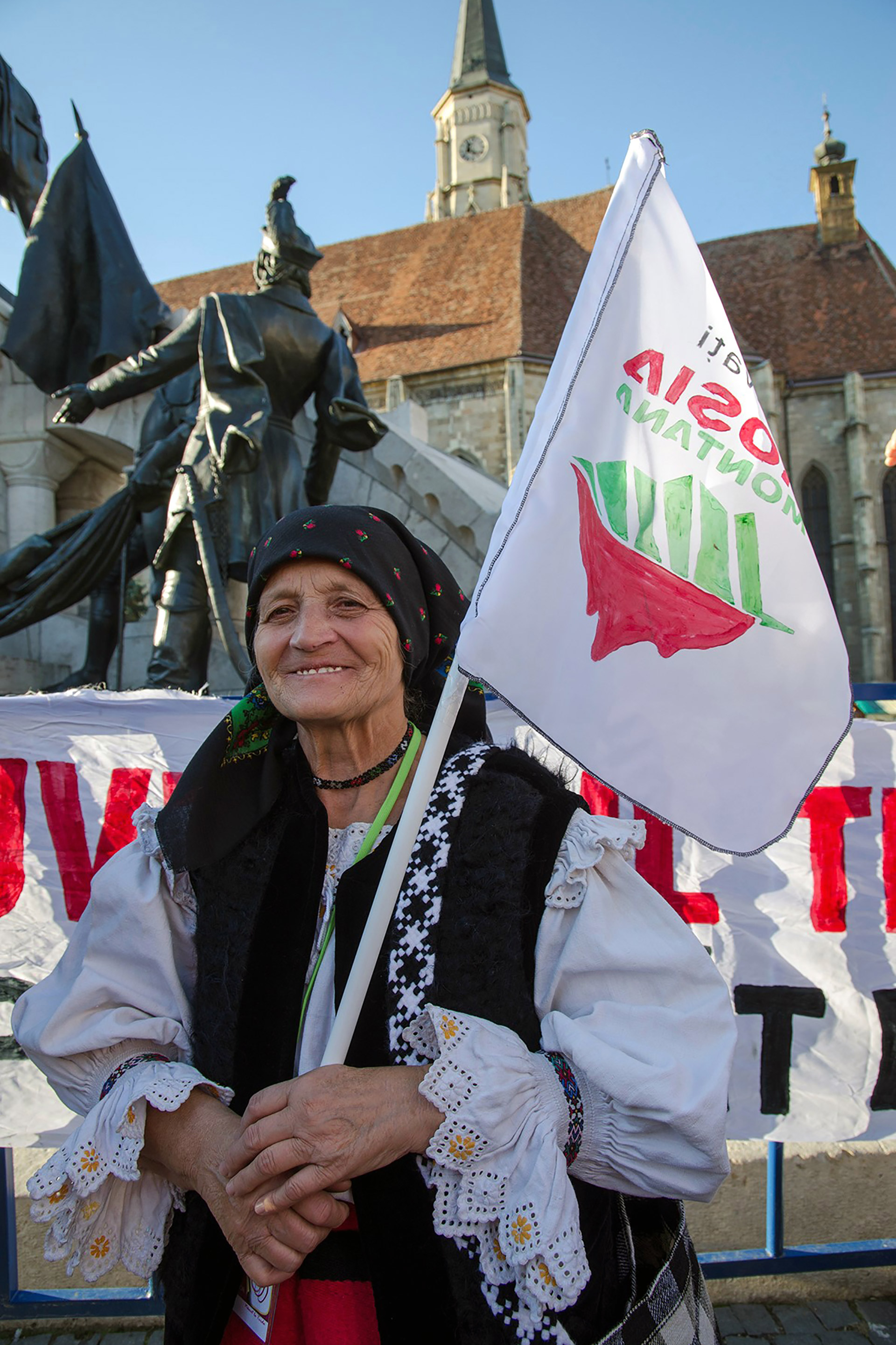 En eldre rumensk dame i folkedrakt står med et flagg med teksten «Salvaţi Roşia Montană»