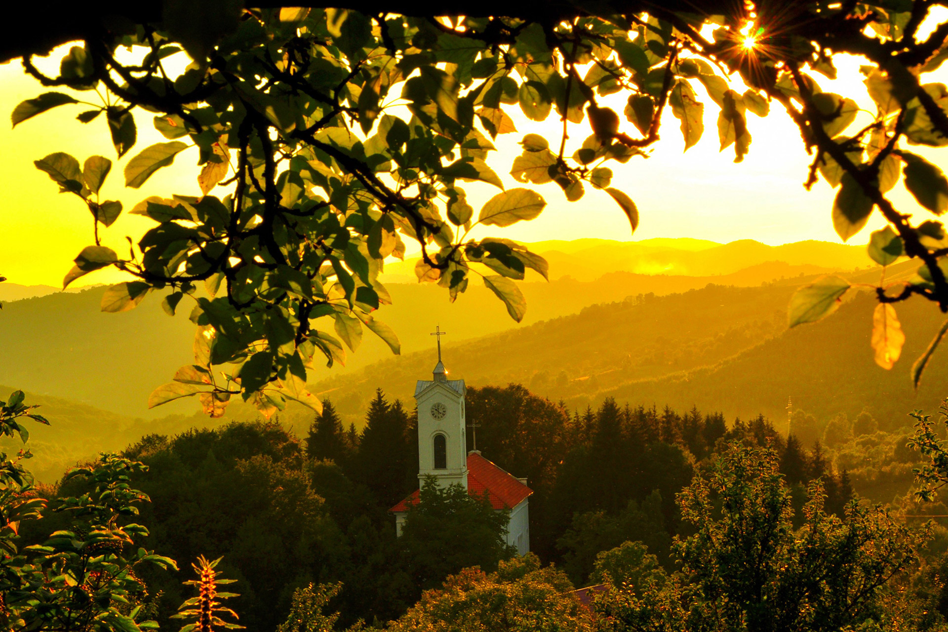 En hvitkalket gammel kirke står på en bakketopp i en grønn dal i gult lys fra solnedgangen. Ved Roşia Montană.