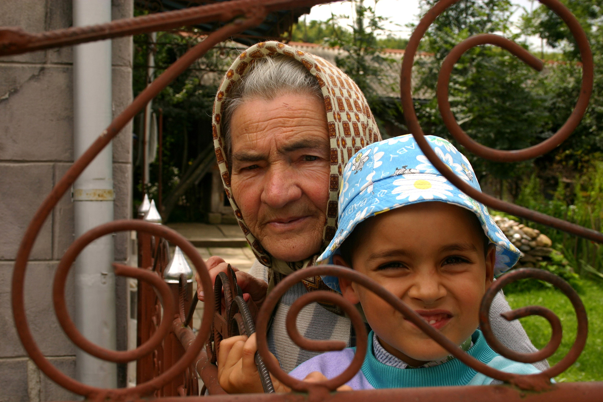 En rumensk bestemor med tradisjonelt skaut står med et barnebarn med solhatt ved en rusten jernport.