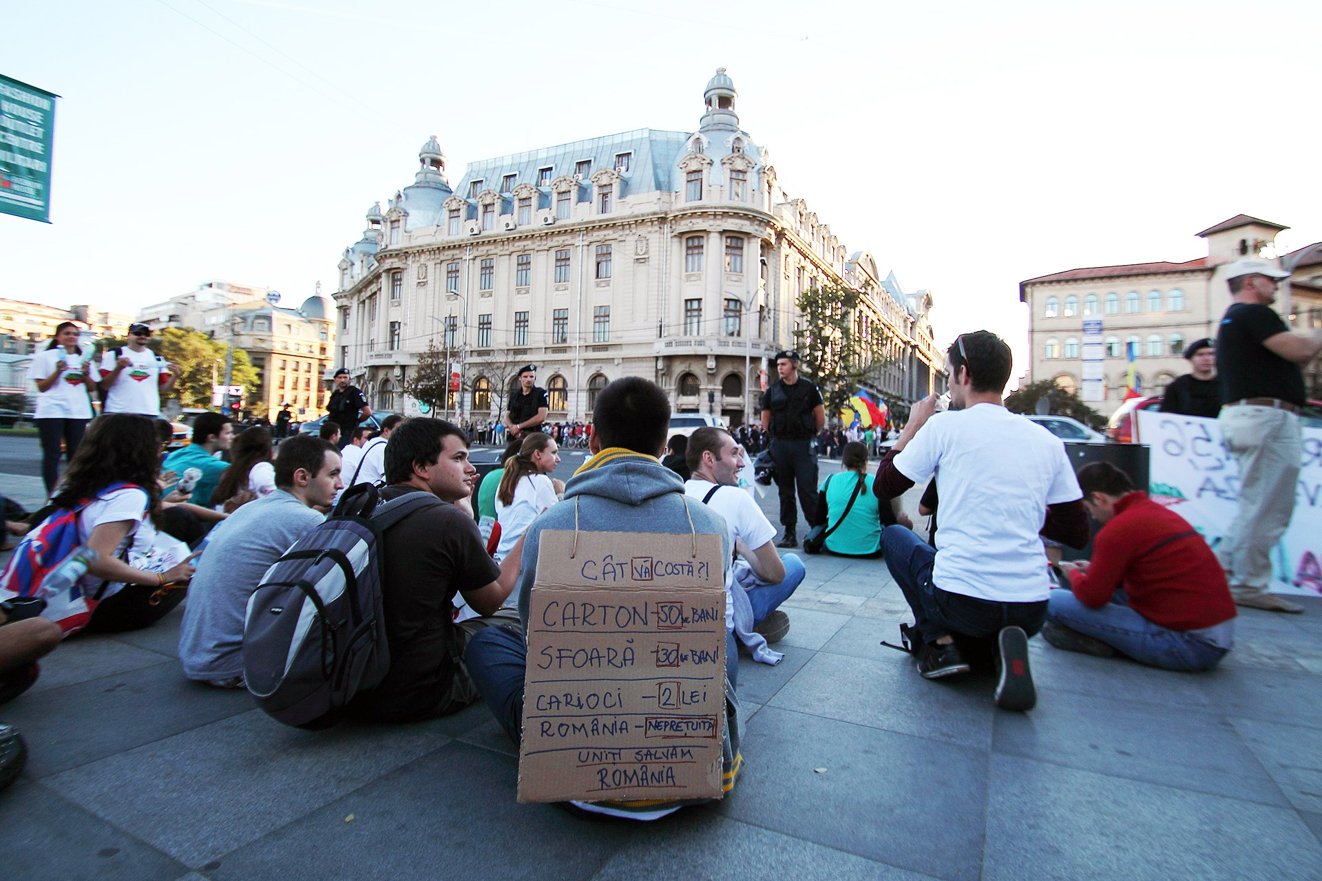 En gruppe demonstranter sitter på bakken med en politimann i bakgrunnen. I forgrunnen har en demonstrant et skilt på ryggen med tekst om at Romania ikke er til salgs. 15. september 2013 ved Piața Universității, Bucuresti.